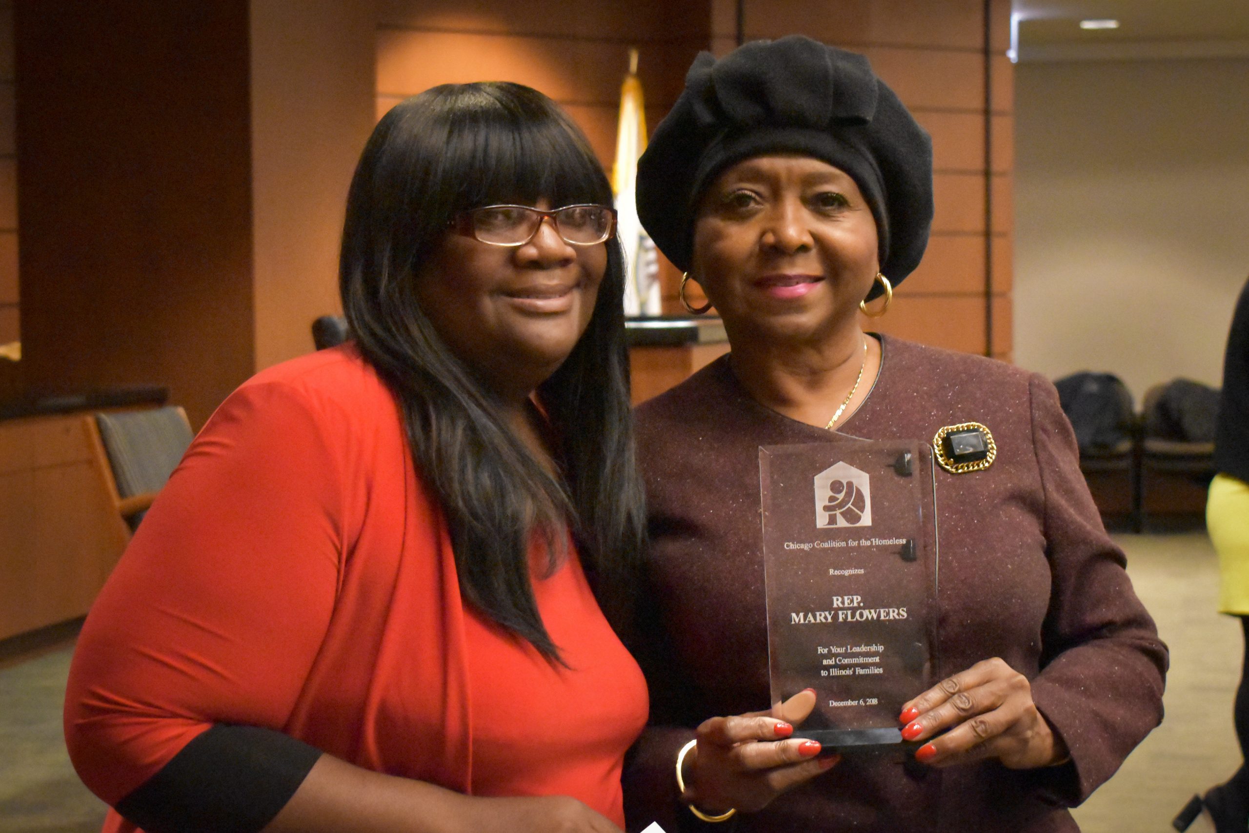 Maxica Williams (Black woman with glasses and straight black hair, wearing a red dress) presents Rep. Mary Flowers (Black woman with gold earrings, black hat, and brown dress) with an award.