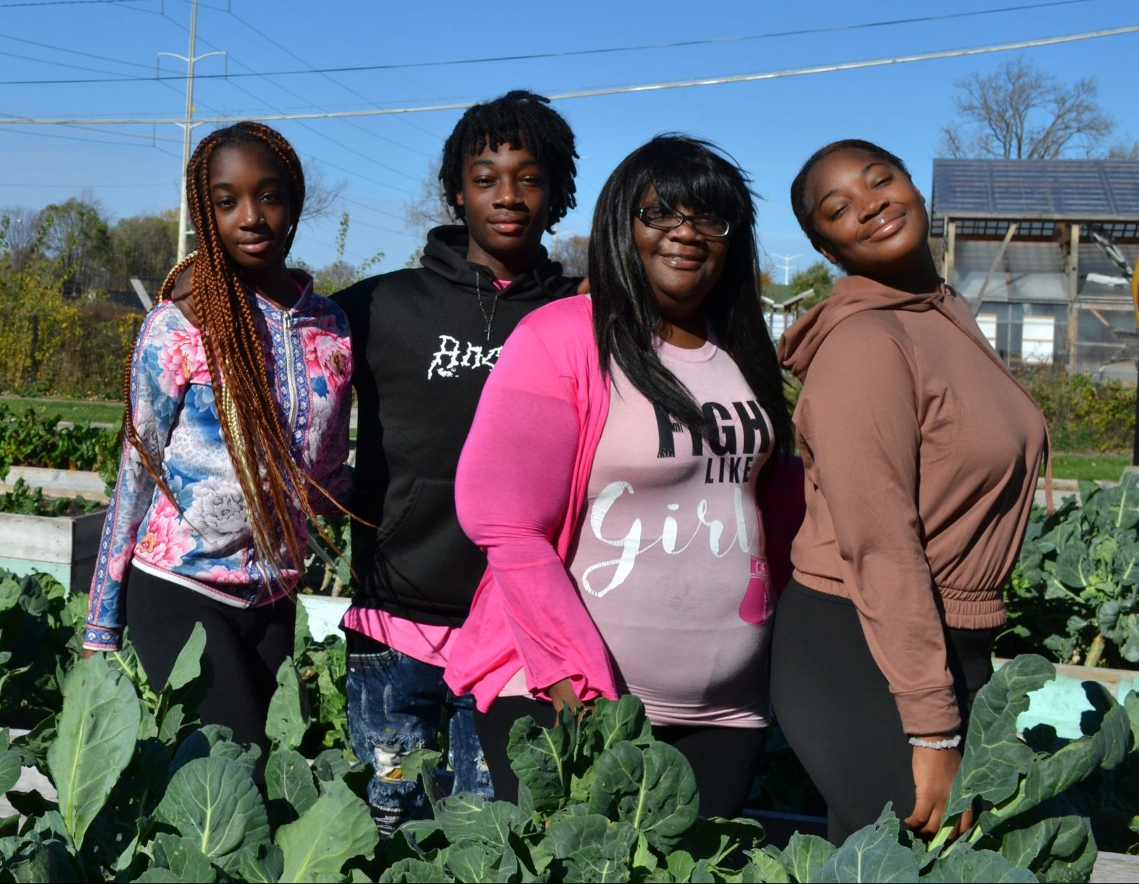 Four members of the Williams family pose in a community garden, surrounded by green leafy vegetables. They include 14-year-old DeSera, 13-year-old DeVon, mother Maxica, and 16-year-old DeNaysa.