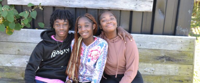 Three Black siblings (ages 13, 14, and 16) pose smiling, sitting on a wooden bench besides a shed.