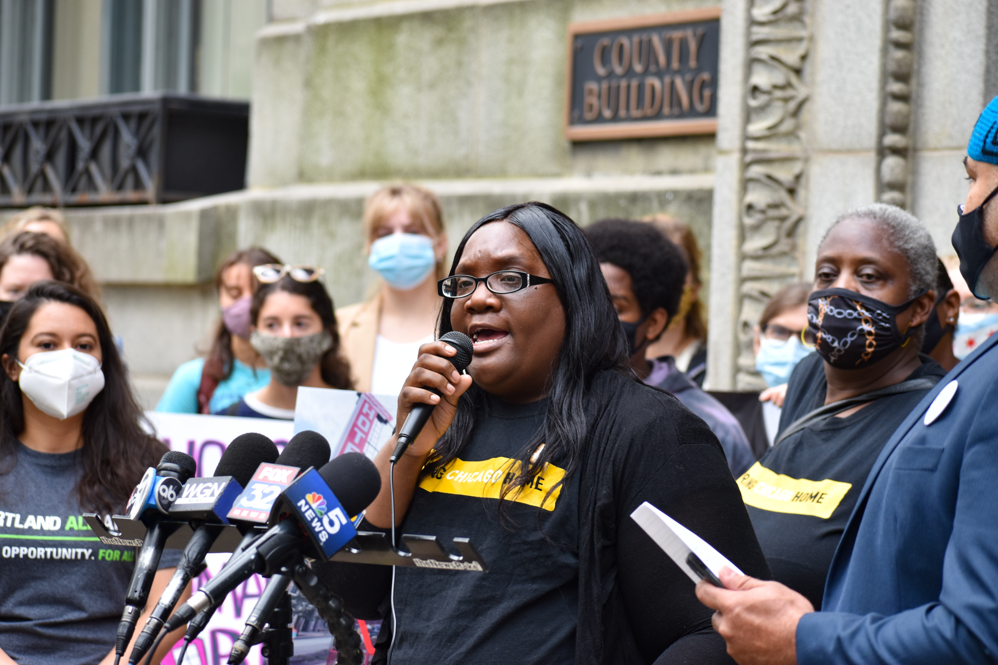 A Black woman speaks to a crowd using a microphone at a press conference in front of Chicago City Hall. She is surrounded by various people of different ages and races, many wearing protective face masks.