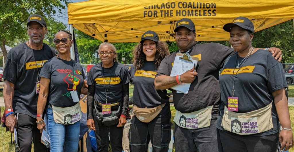 Six Black adults stand smiling in front of a yellow tent with a Chicago Coalition for the Homeless logo. They are wearing black hats and t-shirts with the Bring Chicago Home logo. Several are holding clipboards.