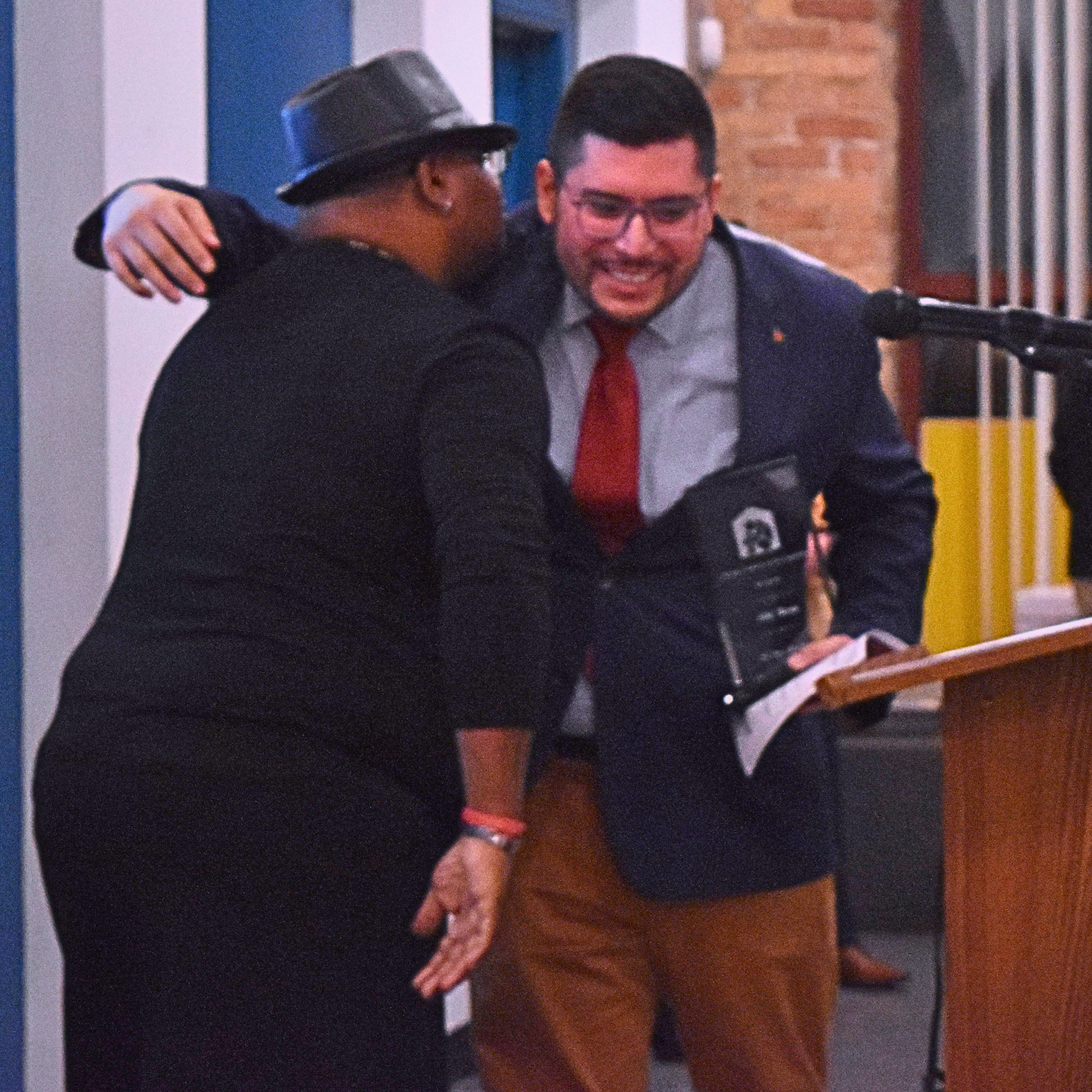 Two men, one Black, one Latino, hug in front of a podium