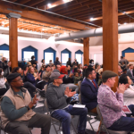 A large group of people sit in folding chairs watching a presentation off camera.