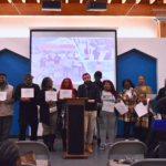 A group of 13 multi-racial, multi-generational people stand side by side behind a podium. Most are holding certificates.