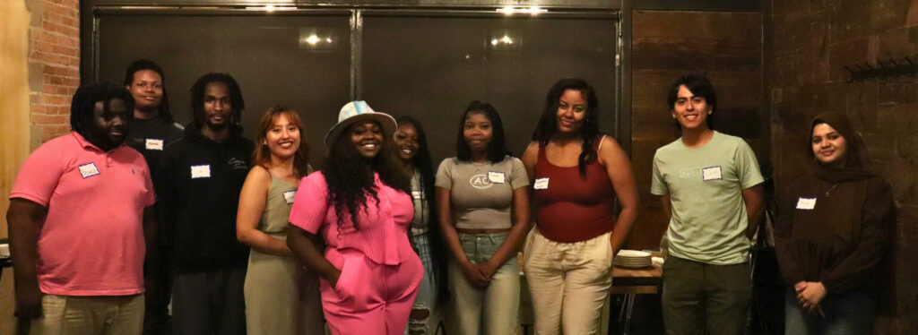 Ten young people of color, aged late teens to mid-20s, stand shoulder to shoulder, posing and smiling for the camera.