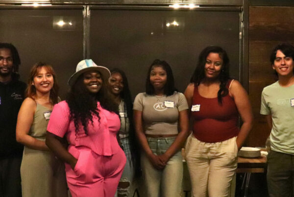 Ten young people of color, aged late teens to mid-20s, stand shoulder to shoulder, posing and smiling for the camera.