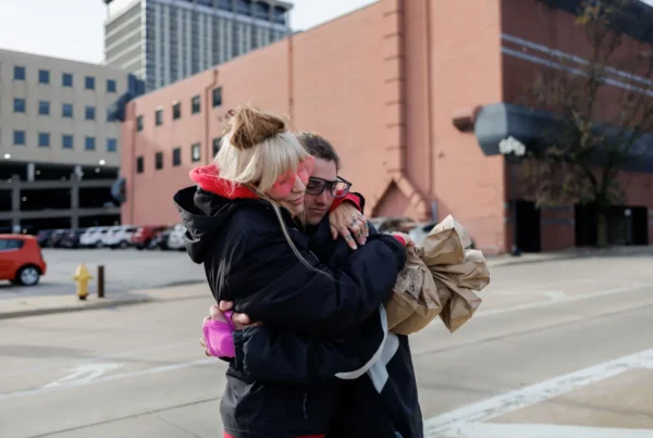 2 people embrace. One is holding many brown lunch bags. They are standing on the sidewalk outside.