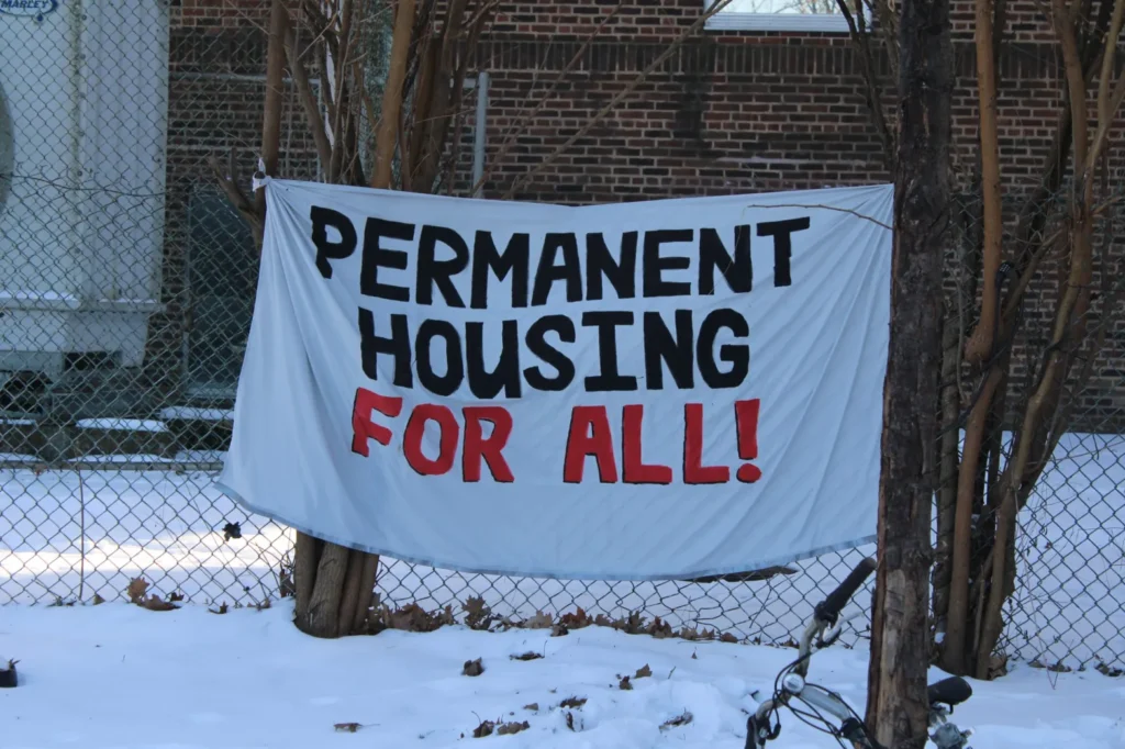 A photography of a sign on a fence that reads "Permanent housing for all". There is snow on the ground.