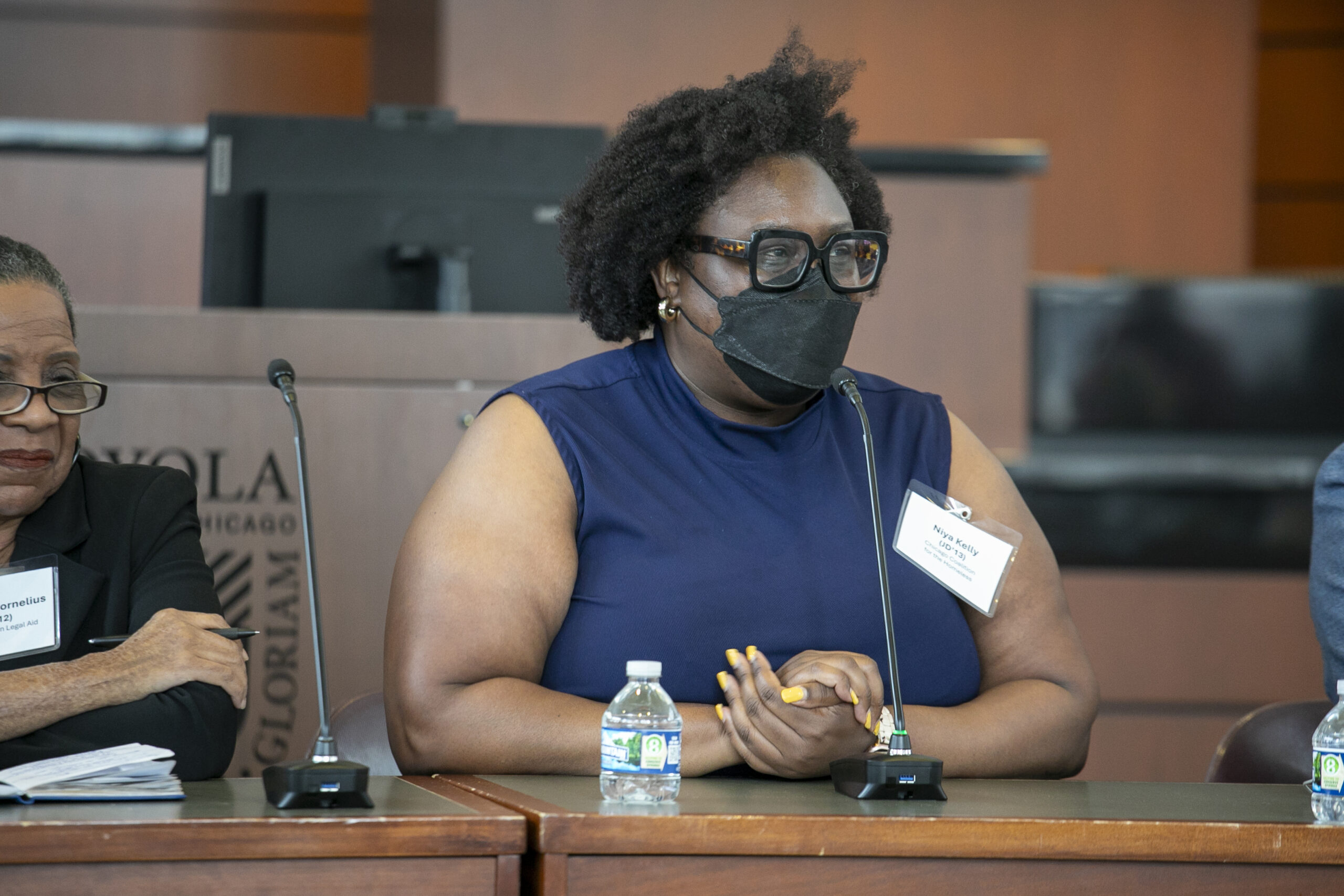 A photograph of a black woman wearing a mask speaking on a panel.