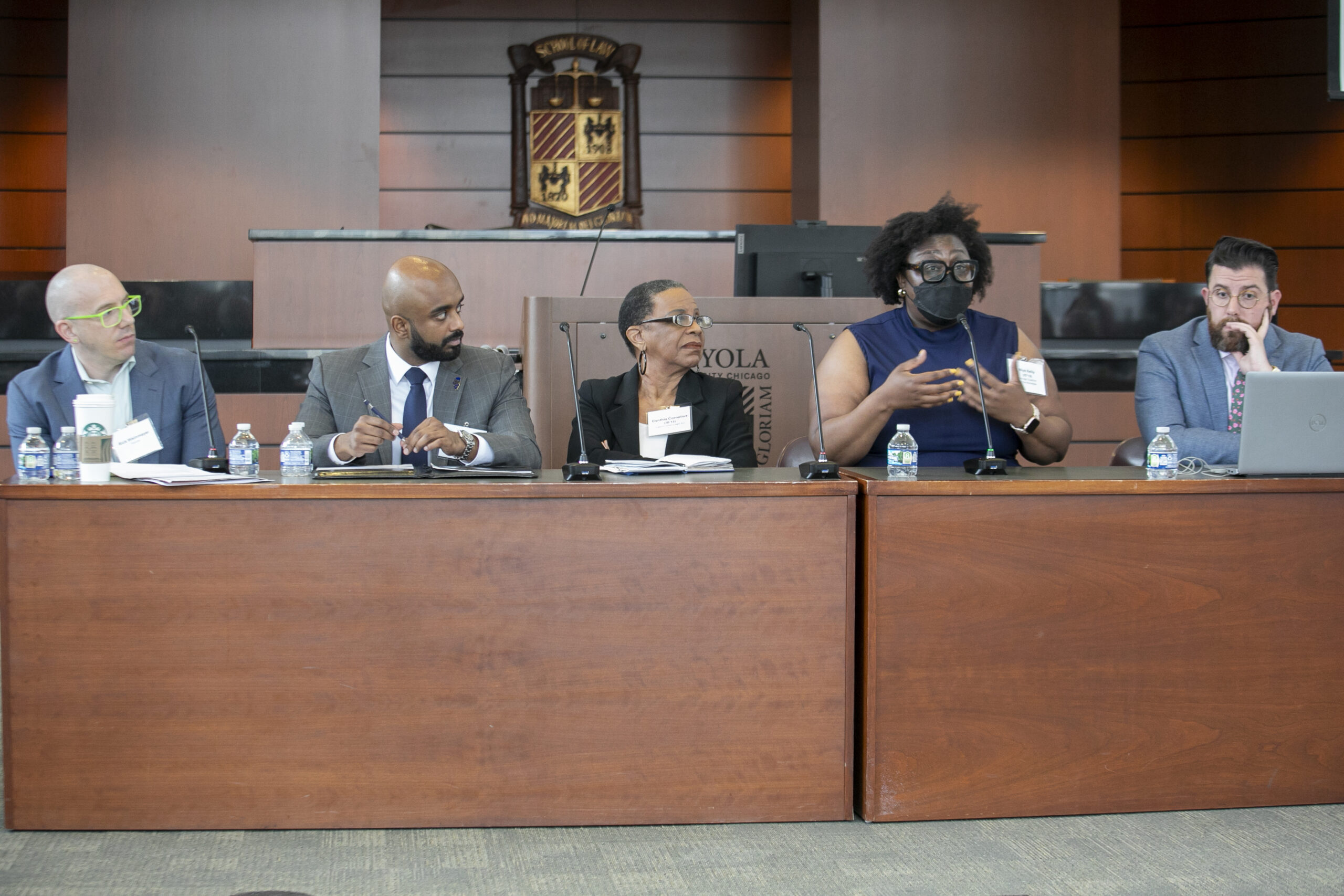 A photograph of panelists at a long wooden table. The Loyola University emblem is in the background.