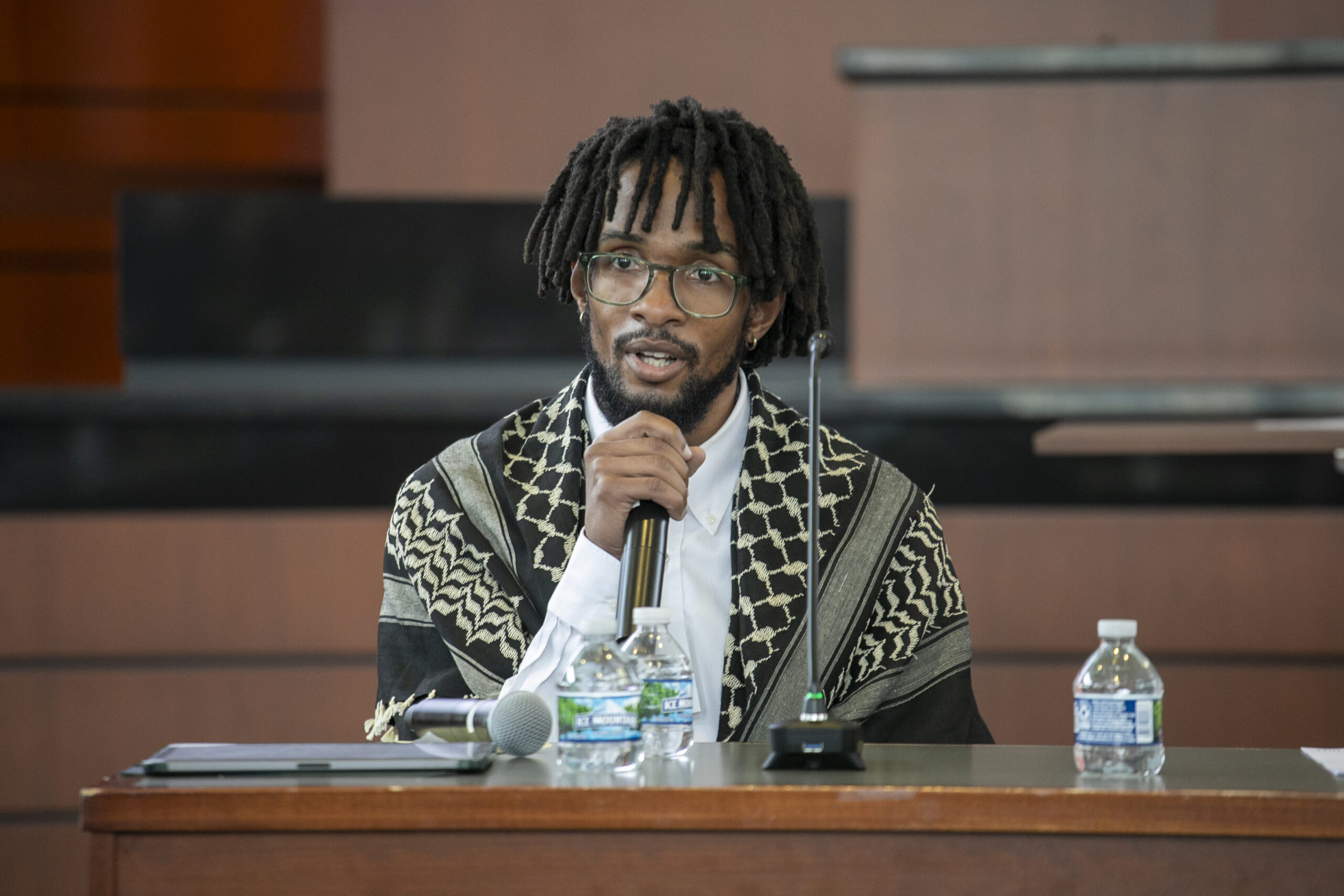 A photograph of a black man with locks wearing a keffiyeh and speaking into a microphone.