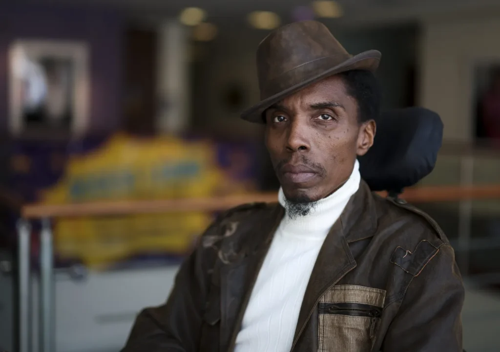 A man in a hat looks at the camera. Her is seated in a wheelchair.