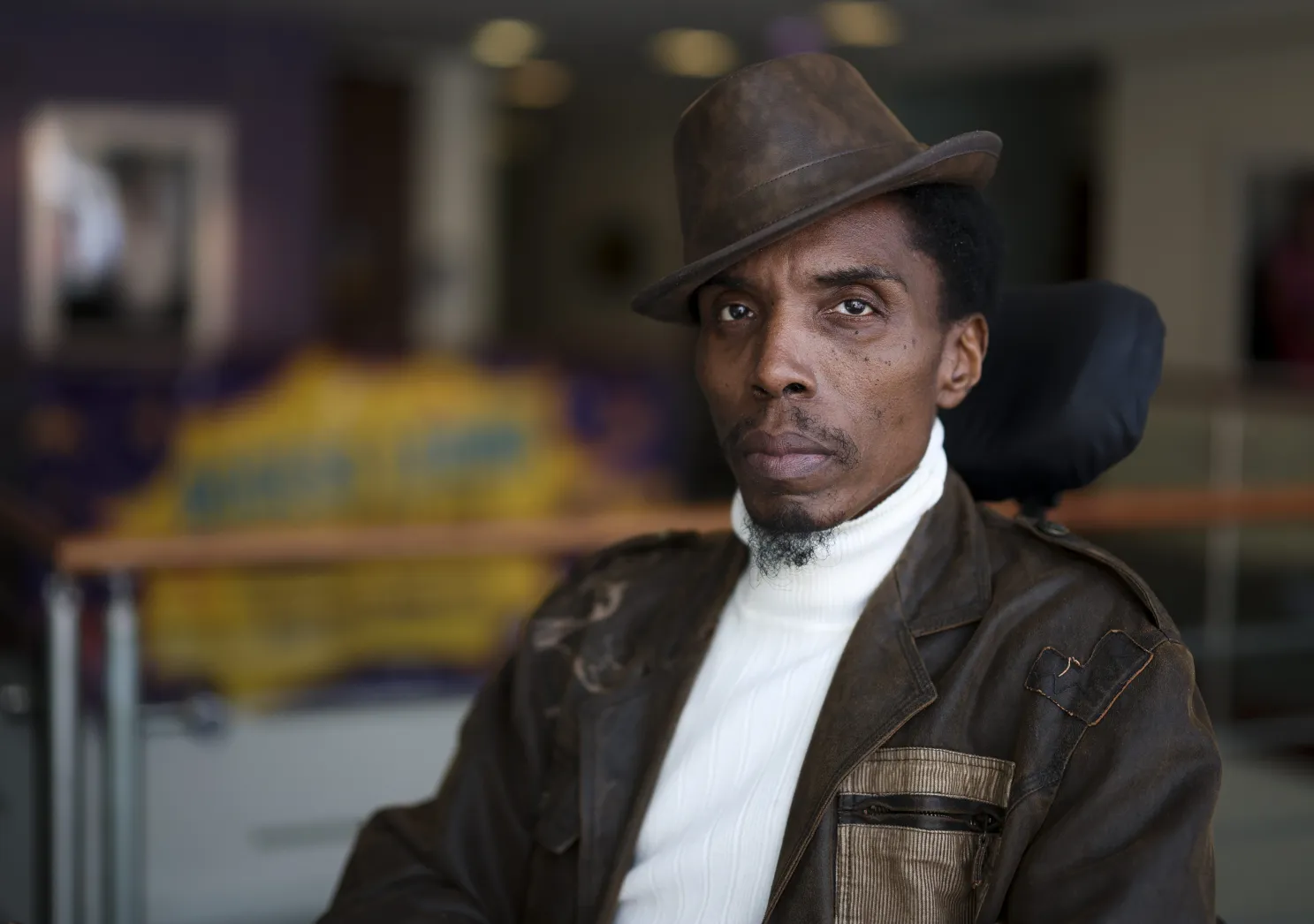A man in a hat looks at the camera. Her is seated in a wheelchair.