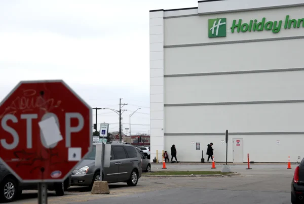 A photograph of a Holiday Inn with people walking. There is a stop sign with stickers and graffiti in the foreground.