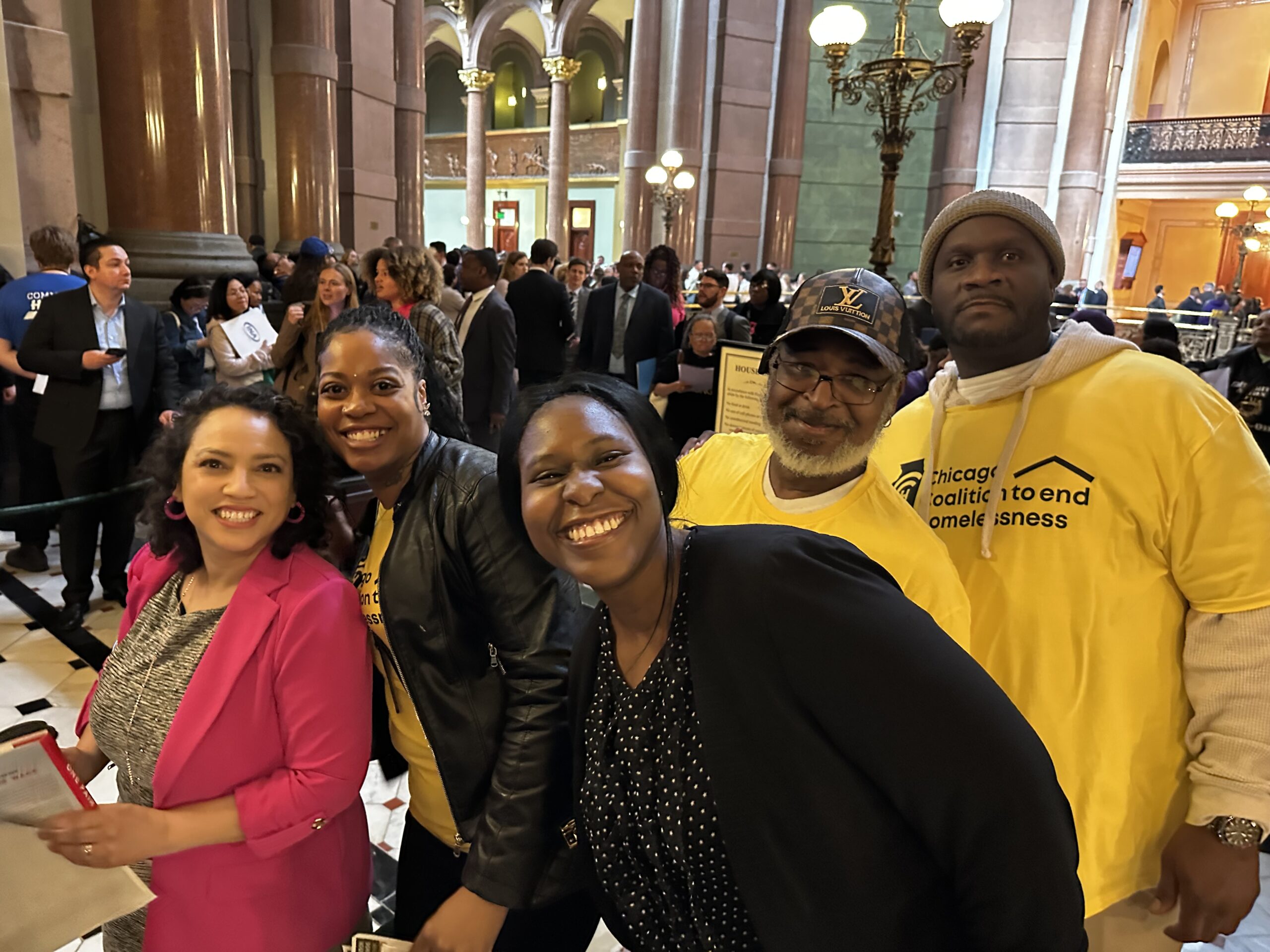 5 people stand together and smile for the camera inside the Illinois State Capitol building. 3 are wearing bright yellow Chicago Coalition to end Homelessness shirts.