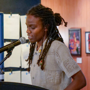 Woman standing at a podium speaking into a microphone. Papers are visible hanging on a wall behind her, as well as wood paneled wall featuring framed art and descriptions.