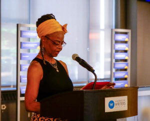 Woman speaking into a microphone at a podium with a American Writers Museum logo on it. Shade-covered windows and signage is visible in the background.