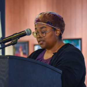 Woman standing at a podium with a microphone reading. Wood paneling and framed images are visible in the background.