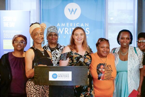 Group of women pose and smile behind a podium with the "American Writers Museum" logo. Logo is also visible on a large poster heind them, along with other signage.