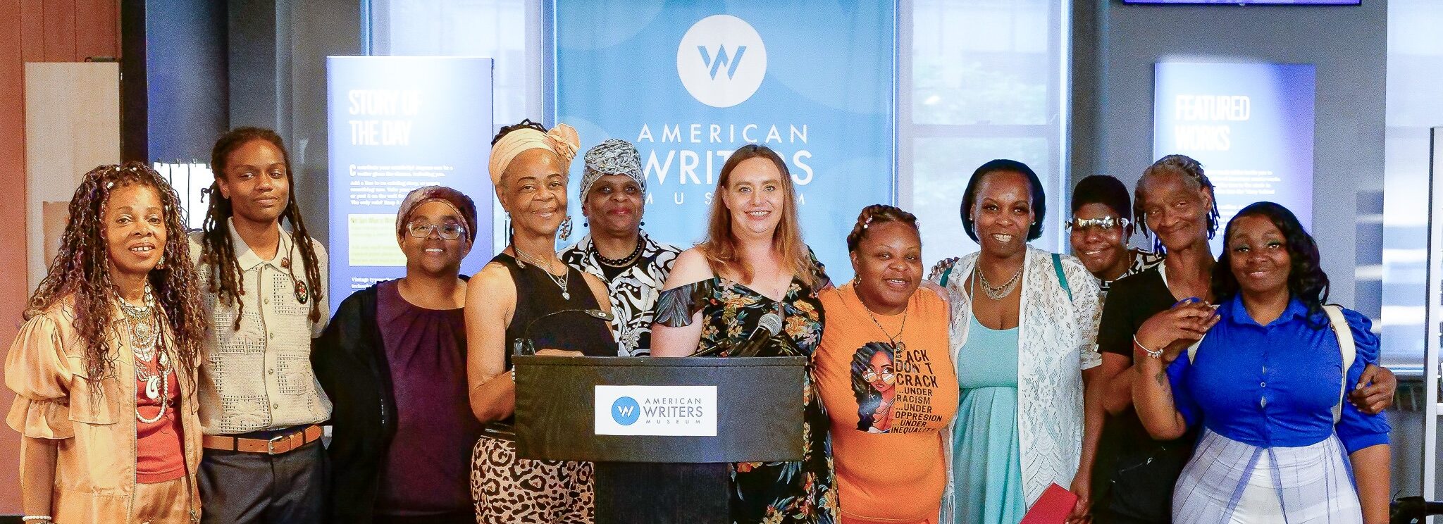Group of women pose and smile behind a podium with the "American Writers Museum" logo. Logo is also visible on a large poster heind them, along with other signage.