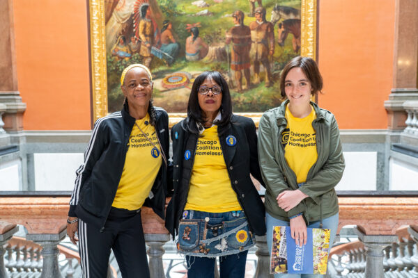 Three people wearing bright yellow shirts stand in front of a painting.