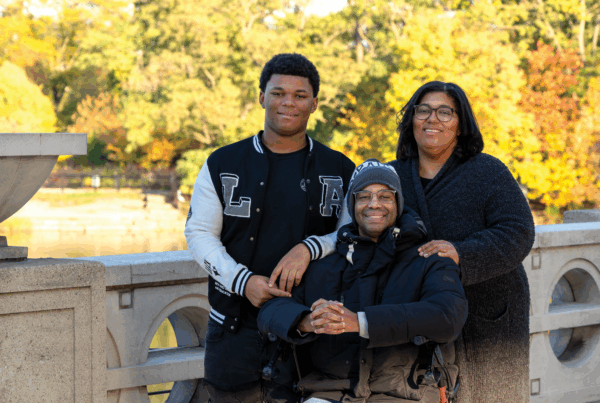 Family of three standing in an outdoor location on a bridge with an fall foliage visible in the background