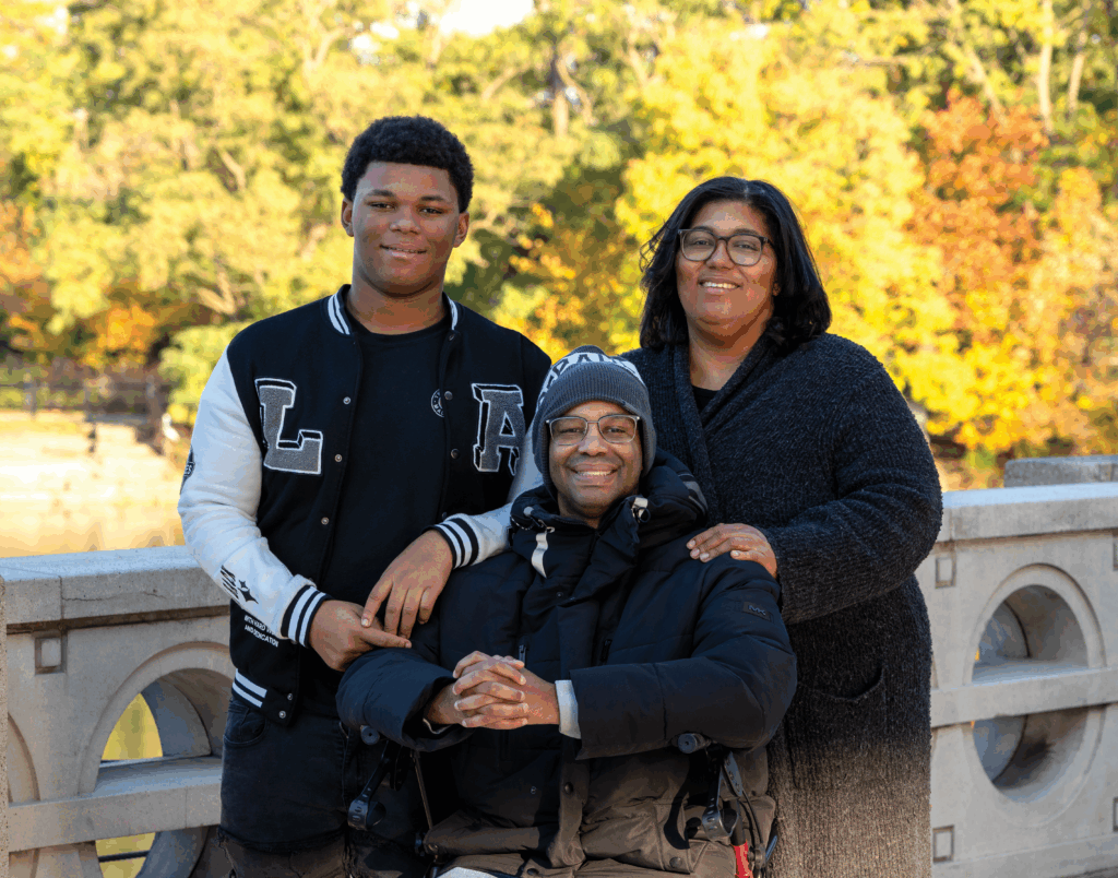 Family of three standing in an outdoor location on a bridge with an fall foliage visible in the background