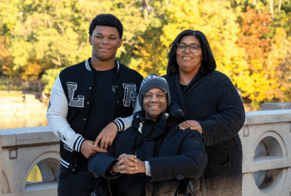Family of three standing in an outdoor location on a bridge with an fall foliage visible in the background