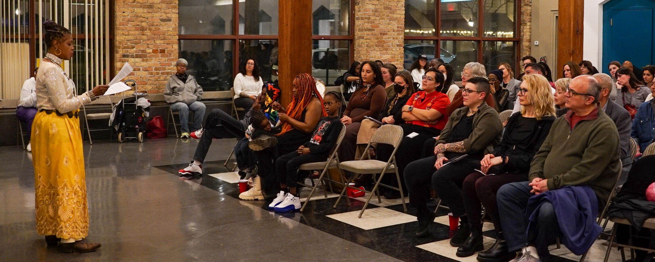 Person in a sparkly white top and yellow skirt stands reading from papers in her hands in front of seated audience listening to her speak.