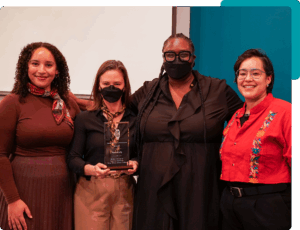Four people smiling and posing for the camera. Two people in the middle are wearing masks, one holds a glass award. 