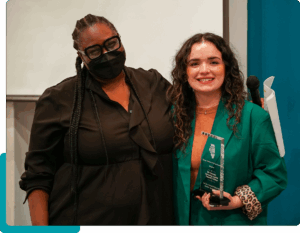 Two people, one wearing a mask both posing and smiling towards the camera. The person on the right holds a glass award.