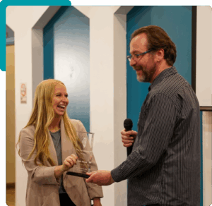 One person handing a glass award plaque to another, both smiling. The person giving the award is holding a microphone.
