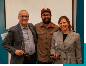 Three people posing and smiling towards the camera. The two on the ends both hold glass award plaques. 