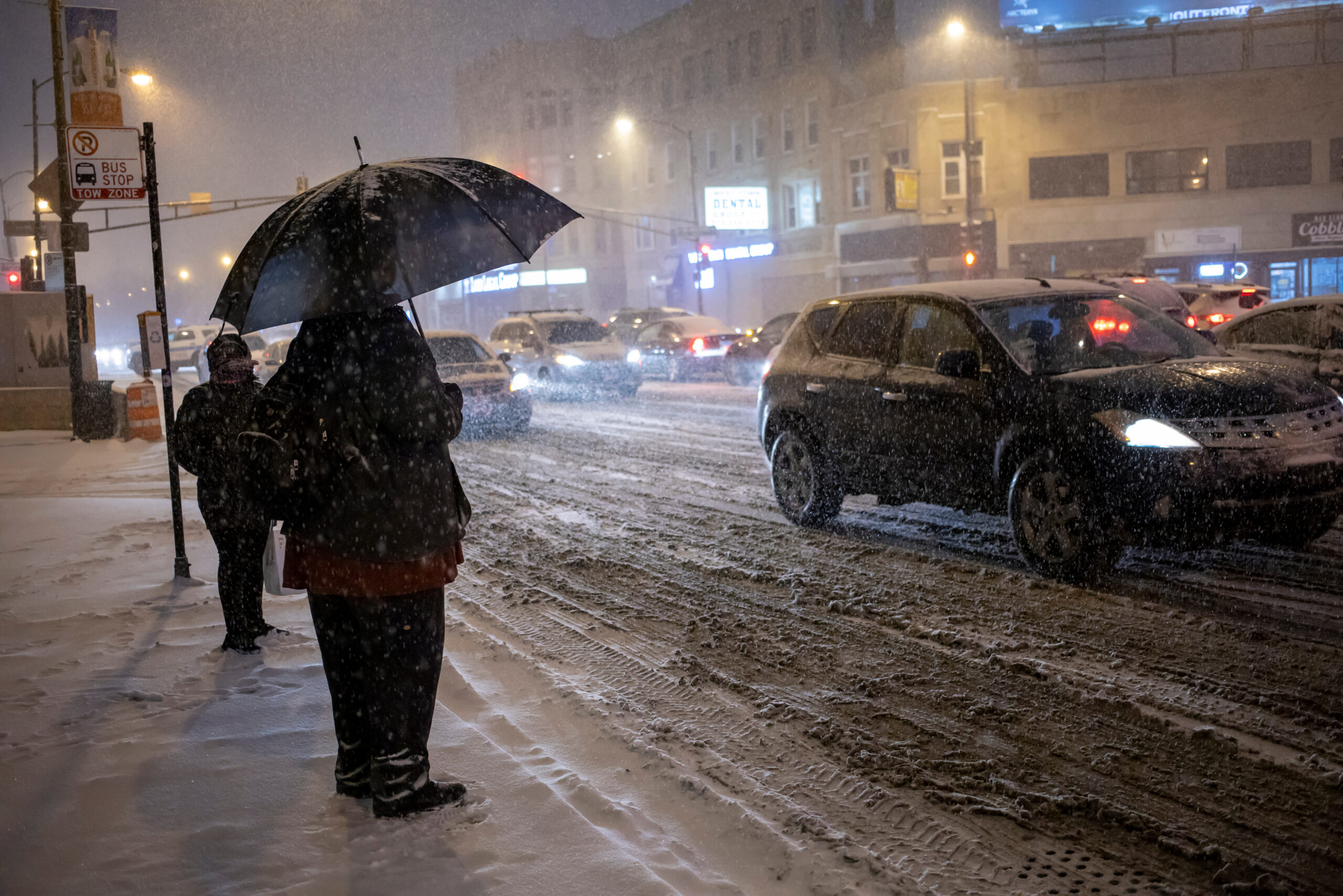 Man is standing outside with umbrella in the extreme cold and snow on a busy Chicago street during a blizzard on a winter night.