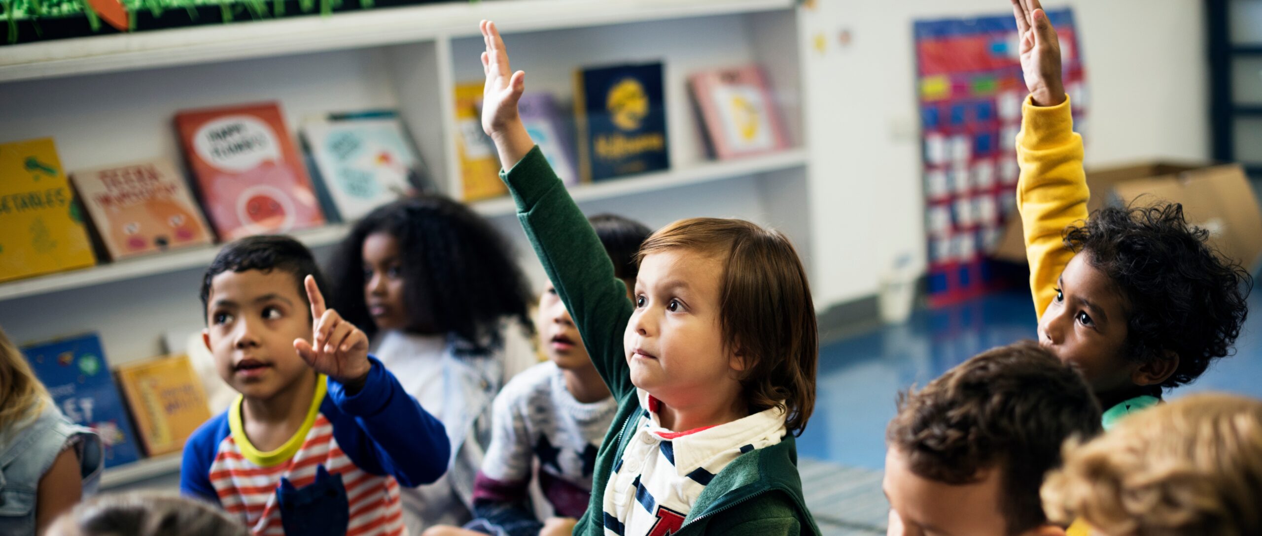 Group of several young students sitting on the floor of a classroom. Three eagerly raise their hands.