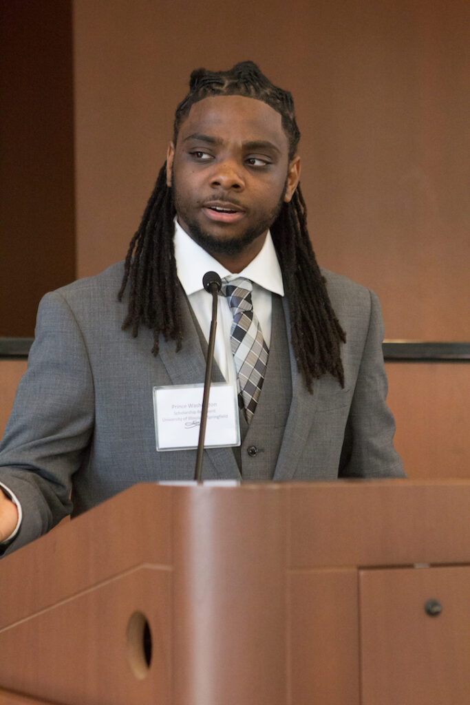 You man stands at a podium speaking into a microphone in a wood-paneled room.