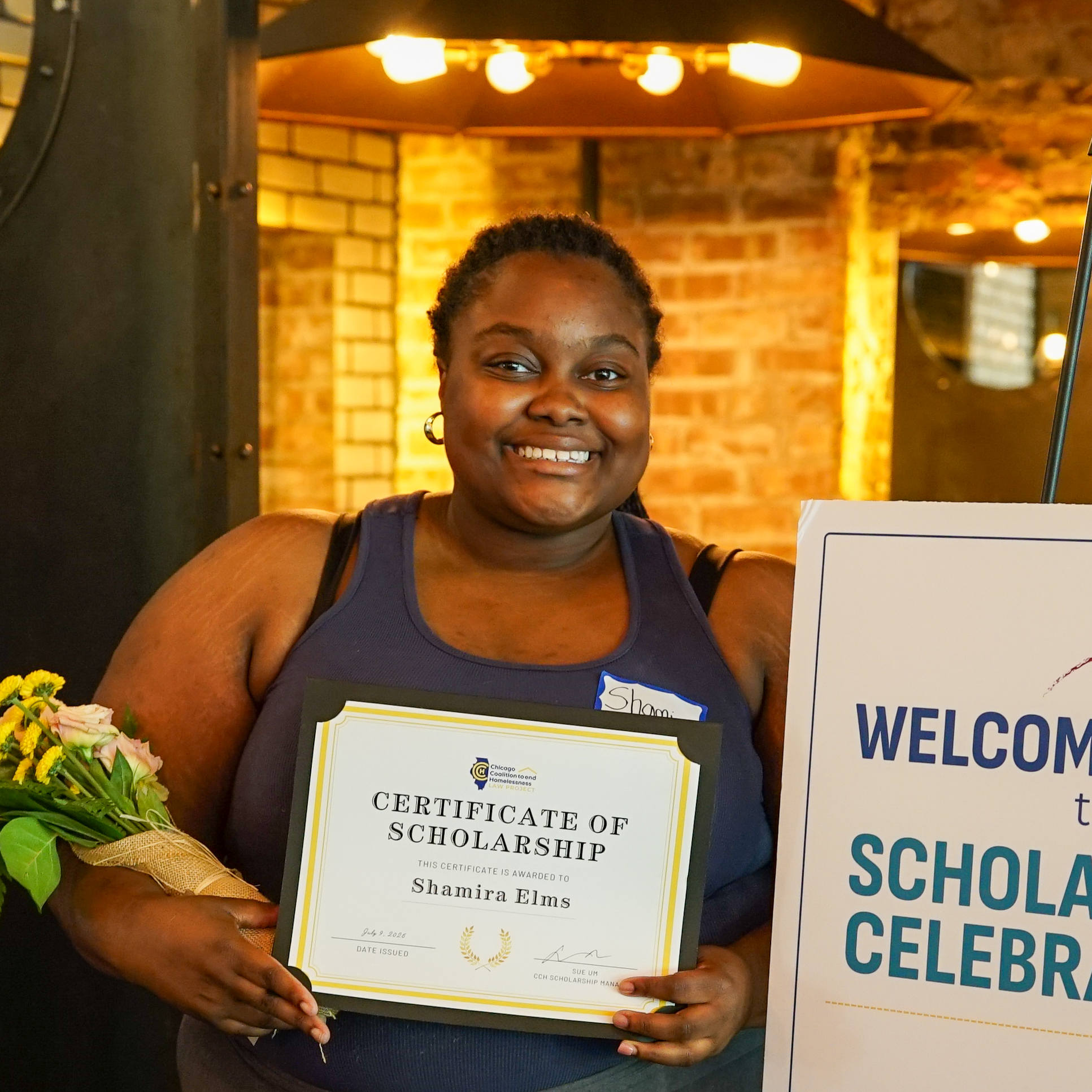 Young woman holding a certificate and flowers in front of a brick wall and overhead lighting