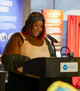Woman stands behind a podium with a microphone reading. Colorful posters and a curtain are visible in the background.